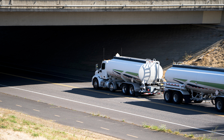 White day cab Big rig semi truck for local distribution transporting two tank semi trailers going under the concrete bridge over the road for fuel delivery to point of destinationの写真素材