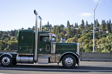 Profile of green classic American pro stylish bonnet big rig semi truck with high vertical exhaust pipes transporting commercial goods and driving on the wide highway with green hill with forestの写真素材