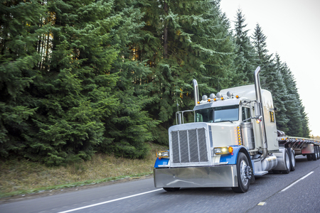 White and blue big rig classic American idol bonnet semi truck with vertical exhaust pipes transporting empty flat bed semi trailer driving on the road with frosted winter evergreen trees on the sideの写真素材