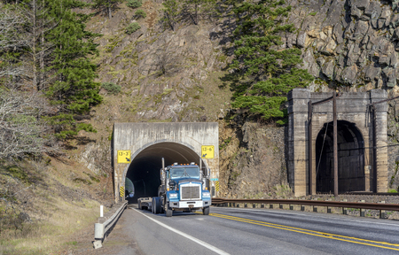 Big rig long haul blue American semi truck transporting commercial cargo on step down semi trailer running through the tunnel on the narrow road along the Columbia River in Columbia River Gorgeの写真素材