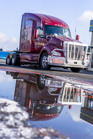 Red big rig semi truck tractor for industrial freight in American logistic system with aluminum bumper grille protector reflecting in puddle of melted snow on winter roadの写真素材