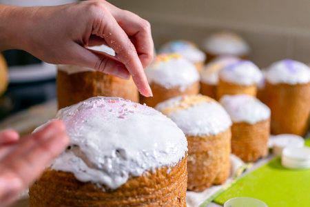 Baker decorates the upper part of Easter cakes covered with white sugar icing by colored granulated...