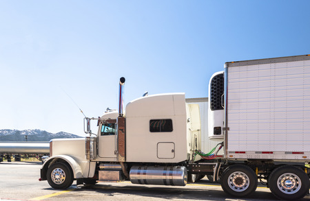 White big rig beautiful classic American powerful semi truck with chrome exhaust pipes with attached refrigerated semi trailer with refrigerator unit on it standing on truck stop parking lot in Utahの写真素材