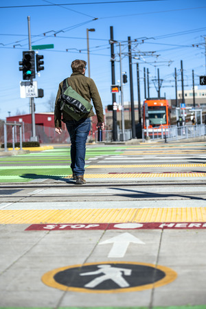 Young man with bag over his shoulder crosses the tram tracks at the intersection of the pedestrian crossing with color graphic marking for pedestrians, goes in direction of the stop of the city tramの写真素材