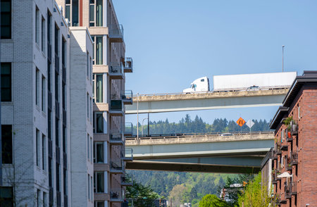 Side view of white big rig long haul semi truck transporting commercial cargo in dry van semi trailer running on the overpass highway between high-rise buildings in urban modern city of Portlandの写真素材