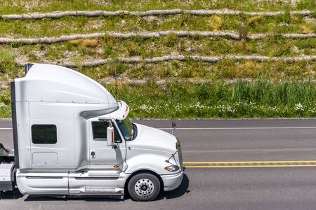 Long haul white big rig semi truck with high cab for truck driver comfort and reduce trailer aerodynamics transporting commercial cargo running on the highway road with green grass on the hillの写真素材