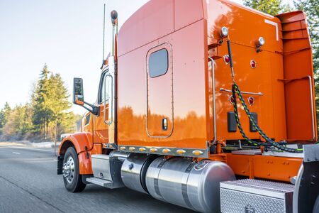 Bright orange classic iconic American big rig long haul professional comfortable powerful semi truck transporting commercial cargo in semi trailer driving on the wide divided multiline highwayの写真素材