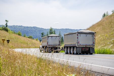 Big rig powerful industrial brown tipper semi tuck tractor with two dump trailers for heavy loads running on the winding road in green trees forest with hills in Columbia Gorge areaの写真素材