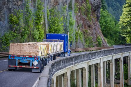 Big rig blue long haul semi truck with two flat bed semi trailers transporting industrial lumber boards running on the winding road with bridge around the mountain rock and the abyss on the sidesの写真素材