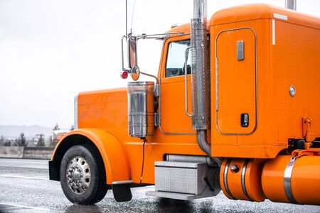Bright orange classic big rig semi truck with vertical exhaust pipes transporting semi trailer running on wide multiline wet road in raining weatherの写真素材