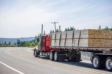 Big rig red classic powerful long haul semi truck with flat bed semi trailer transporting tightened industrial lumber boards running on the turning winding road with hills and trees in sunny dayの写真素材