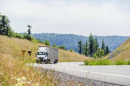 Big rig white day cab semi truck with roof spoiler transporting huge covered bulk semi trailer moving uphill on the turning winding road with yellow hills and green treesの写真素材