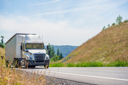 Big rig white day cab semi truck with roof spoiler transporting huge covered bulk semi trailer...