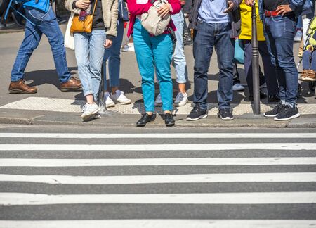 A number of people in various clothes lined up at a pedestrian crossing waiting for a traffic light to go across the street to the other side of the street.の写真素材