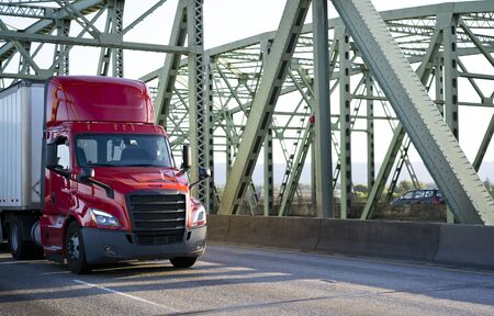 Big rig bright red day cab powerful bonnet semi truck transporting commercial cargo in dry van semi trailer with aerodynamic skirt driving on the metal truss Columbia River Interstate drawbridge Big rig bright red day cab powerful bonnet semi truck transporting commercial cargo in dry van semi trailer with aerodynamic skirt driving on the metal truss Columbia River Interstate drawbridge