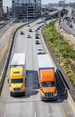 Two different models of big rig orange and yellow semi trucks with dry van and refrigerator semi trailers running in front of another traffic on the road with overpass intersections in city limitの写真素材