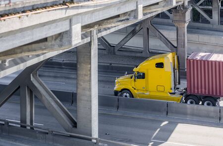 Big rig powerful Yellow long haul industrial grade semi truck transporting commercial goods in container on flat bed semi trailer moving on the divided interstate highway road under the bridgeの写真素材