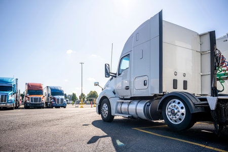 Long haul big rig white semi truck tractor stand on truck stop parking lot across another semi trucks standing in row for truck drivers rest waiting for the continuation of the cargo delivery routeの写真素材