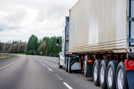 Big rig heavy-duty long haul diesel semi truck with additional axles transporting commercial cargo loaded container on semi trailer moving on the wide highway with marked lines with stormy cloudsの写真素材