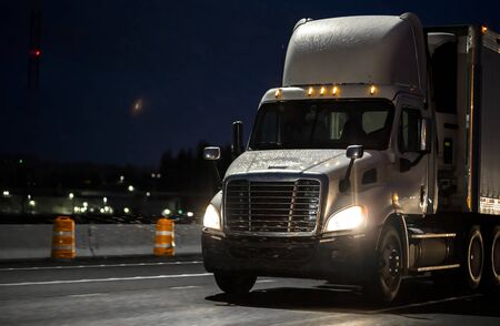 Big rig day cab heavy-duty diesel semi truck with roof spoiler transporting commercial cargo in semi trailer running on the wet raining highway road at dark night time with turned on headlightの写真素材