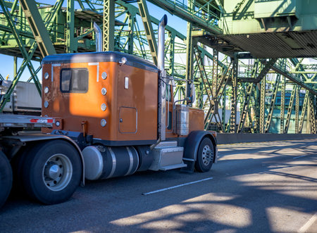 Classic American idol big rig efficient diesel semi truck with chrome accessories and extra light transporting heavy equipment on flat bed semi trailer running on Columbia River Interstate Drawbridgeの写真素材