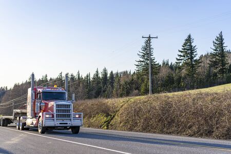 Industrial transportation Red big rig diesel semi truck with large grille guard transporting empty step down semi trailer for carry oversized loads running on the road with field hill on the sideの写真素材