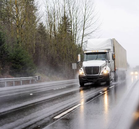 Big rig white diesel semi truck with turned on headlights transporting container with commercial cargo running on the wet multiline highway at raining weather with reflection on the road surfaceの写真素材