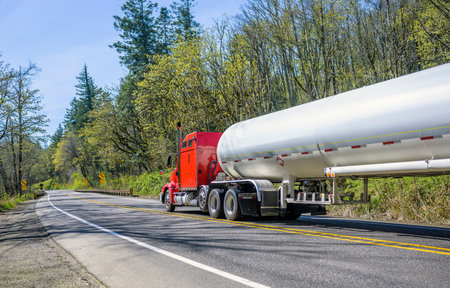 Red big rig classic long haul semi truck transporting commercial flammable cargo in tank semi trailer for for transportation of liquefied gas and other chemicals running on the road in spring forestの写真素材