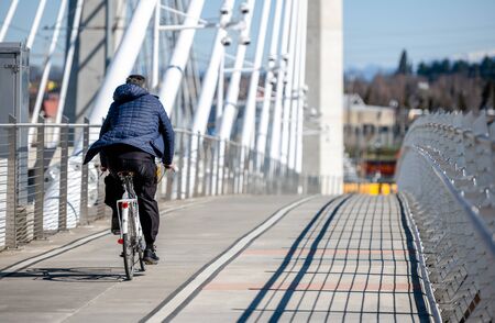 An elderly man on a bike pedals a bicycle on the Tilikum Crossing Bridge preferring an active healthy lifestyle using cycling ride and cycle as an alternative environmentally friendly transportationの写真素材