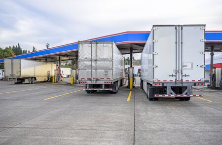 Different long haul diesel Big rigs semi trucks with semi trailers standing on truck stop fuel station for refueling and continue delivering route in accordance with the log book scheduleの写真素材