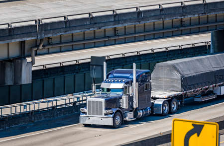Dark blue classic industrial diesel big rig semi truck tractor transporting covered commercial cargo on flat bed semi trailer running on multilevel overpass highway road intersectionの写真素材