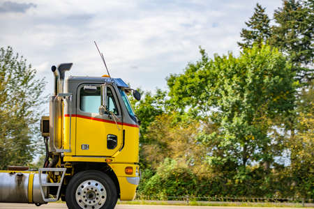 Compact powerful cab over engine big rig yellow semi truck tractor transporting fuel in two industrial tank semi trailers driving on the divided highway road with trees on the sideの写真素材