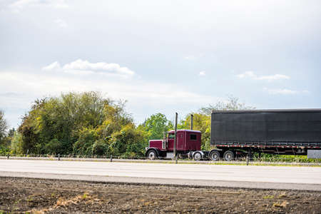 Big rig burgundy classic American industrial semi truck tractor with sleeper cab compartment for truck driver rest transporting cargo in dry van semi trailer driving on the highway road with treesの写真素材