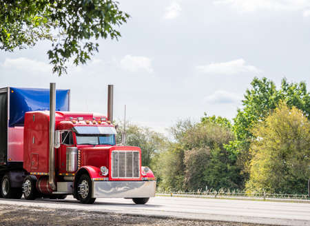 Big rig red classic American industrial semi truck tractor with sleeper cab compartment for truck driver rest transporting cargo in dry van semi trailer driving on the highway road with treesの写真素材