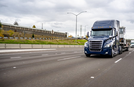 Dark blue industrial big rig semi truck tractor with high cab transporting industrial truck on step down semi trailer running on the wide multiline highway road in front of another trafficの写真素材