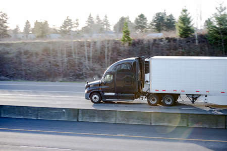 Powerful Big rig industrial classic black semi truck tractor transporting frozen commercial cargo in refrigerated semi trailer running for delivery on the wide divided highway road with sun lightの写真素材