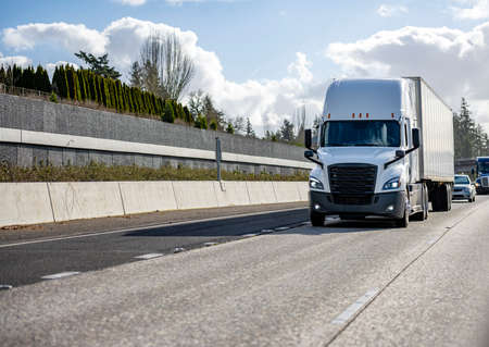 Classic big rig white semi truck tractor with sleeping compartment transporting commercial  cargo in dry van semi trailer moving on the highway road intersection with one way traffic directionの写真素材