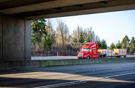 Classic big rig industrial red semi truck tractor with high roof cab and sleeping compartment transporting lumber cargo on step down semi trailer running on the wide highway under the bridgeの写真素材