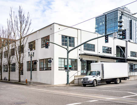Commercial compact day cab big rig semi truck with long box trailer for local freight and deliveries standing on the urban city street crossroad with multilevel buildings and traffic lightの写真素材