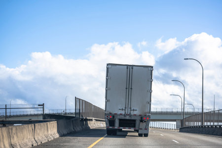 Big rig industrial powerful semi truck transporting commercial cargo in dry van semi trailer climbing on the inclined overpass highway road intersection with bridge and road junction aheadの写真素材