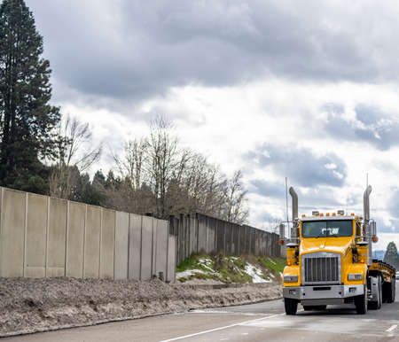 Classic yellow American day cab big rig semi truck transporting empty step down semi trailer for oversized loads running on the winter highway road with concrete fenceの写真素材