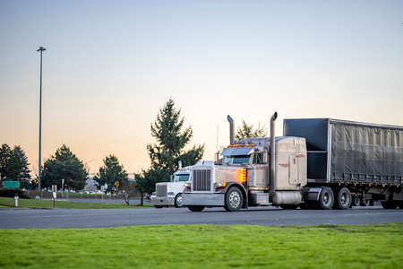 Two classic American Idol big rigs semi trucks with shiny chrome parts and high vertical exhaust pipes and dry van semi trailers standing on the parking lot for rest according to the scheduleの写真素材