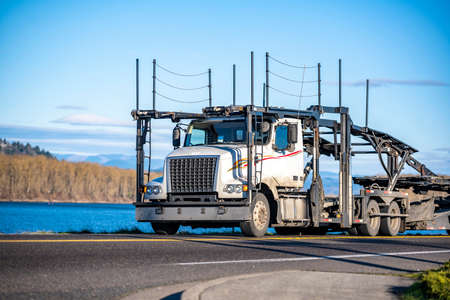 Classic Big rig industrial car hauler semi truck with empty double deck hydraulic modular semi trailers running on the narrow road along the river to warehouse for pick up next cars loadの写真素材