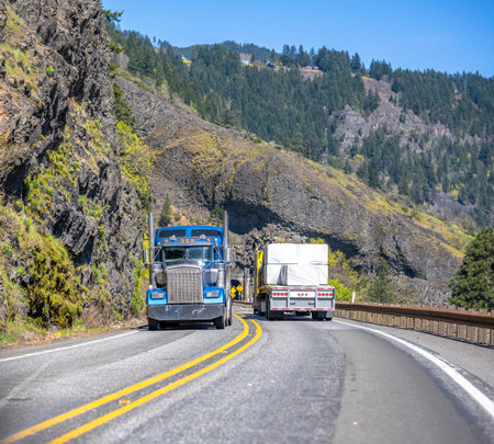 Different classic loaded big rigs semi trucks with semi trailers moving in opposite directions on the narrow winding road with tunnel and rock on the side at national Columbia Gorge Areaの写真素材