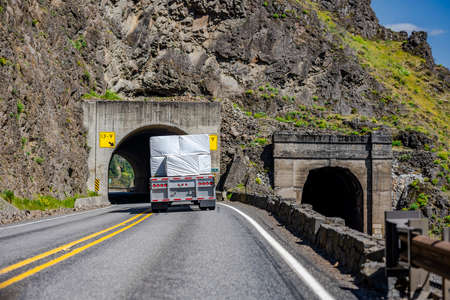 Powerful classic big rig semi truck transporting flat bed semi trailer loaded with covered lumber cargo turning on the winding highway road through a tunnel in the rock at Columbia River Gorgeの写真素材