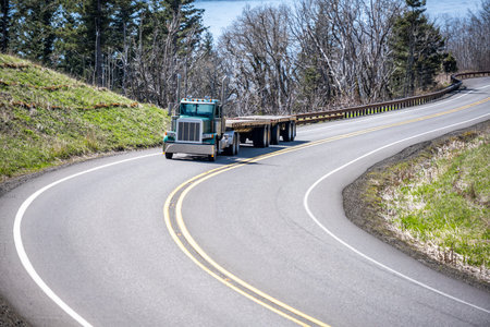 Powerful industrial day cab dark green classic big rig semi truck tractor with two empty flat bed semi trailers running on the winding mountain road at Columbia Gorge to warehouse for next loadの写真素材