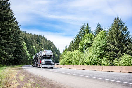 White compact big rig industrial car hauler semi truck transporting cars on the hydraulic modular two level semi trailer running on curving highway road with green mountain forest in Columbia Gorgeの写真素材