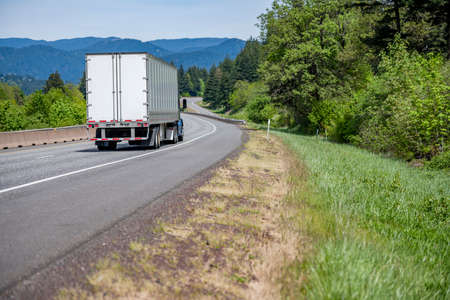 Blue big rig industrial semi truck tractor with roof spoiler transporting cargo in dry van semi trailer running on curving highway road with green forest trees and mountain in Columbia Gorgeの写真素材