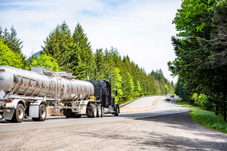 Black classic big rig industrial semi truck tractor transporting liquid cargo in stainless steel tank semi trailer running on curving highway road with green mountain forest in Columbia Gorgeの写真素材