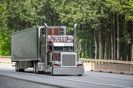 Brown big rig semi truck tractor with vertical chrome pipes transporting cargo in polished corrugated aluminum refrigerator semi trailer running on the green forest highway road in Columbia Gorgeの写真素材